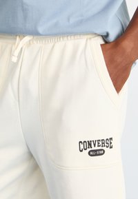 Pantalons de jogging crème avec cordon de serrage, arborant le logo "Converse All-Star" en noir sur le côté gauche, texture lisse et poche latérale.