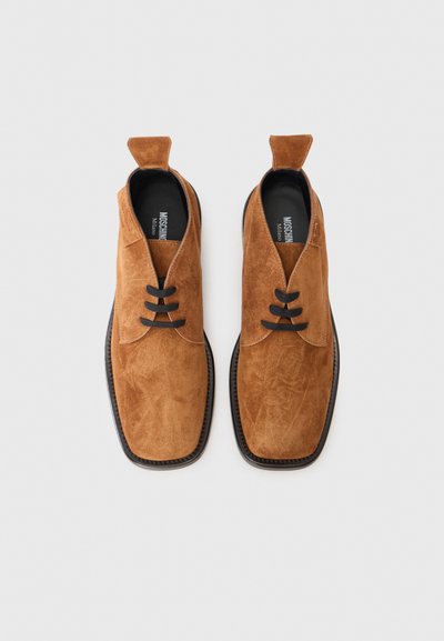 Zapatos de ante marrón con una textura suave, punta redonda y cordones negros planos. Presentan una lengüeta distintiva en el talón y una suela oscura.