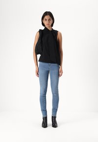 Svart ärmlös topp med knutdetalj, matchad med ljusblå skinny jeans och svarta ankelboots. Modellen håller en strukturerad svart väska.