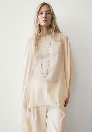 Donna con capelli biondi lunghi, indossa una maglia beige trasparente, pantaloni larghi color crema e una lunga collana d'argento, su sfondo bianco semplice.