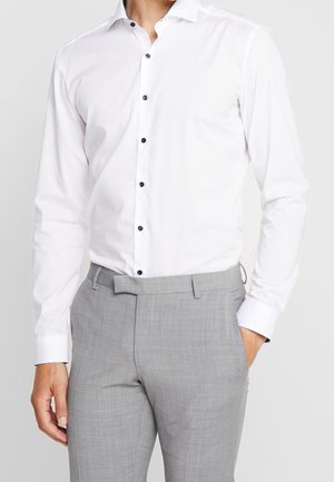Mann trägt ein weißes langärmliges Hemd mit dunklen Knöpfen und hellgraue maßgeschneiderte Hose, Hand in der Tasche.