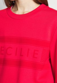 Röd sweatshirt med rund hals, texturerad horisontell rand och upphöjd typografi. Slät yta med ett mönster på ena sidan.