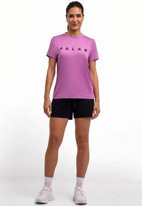Lila Sportshirt mit schwarzem "FALKE"-Schriftzug, kombiniert mit schwarzen Shorts. Das Modell trägt weiße Sneakers und Knöchelsocken. Schlichtes Design.