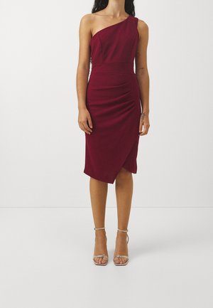 Woman wearing a sleeveless, one-shoulder, knee-length burgundy dress with an asymmetrical hem and silver ankle-strap heels.