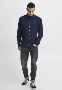 Camicia blu navy con taschino, maniche lunghe, bottoni bianchi, abbinata a jeans grigi e sneakers nere con dettagli bianchi.