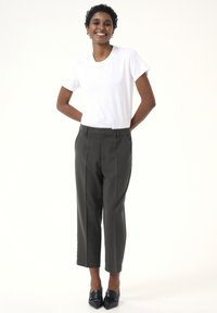 T-shirt blanc à manches courtes associé à un pantalon tailleur vert foncé, avec des plis avant et une coupe droite, complété par des chaussures noires à bouts pointus.