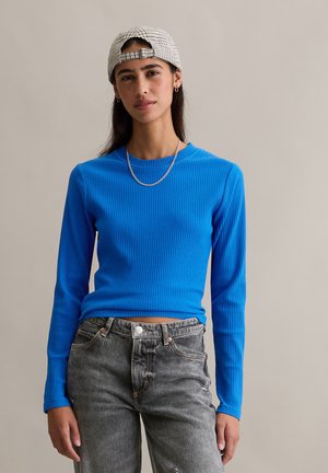 Blaues, geripptes Langarmshirt kombiniert mit grauen, abgenutzten Jeans. Das Outfit wird ergänzt durch eine helle karierte Mütze und eine silberne Kettenhalskette.