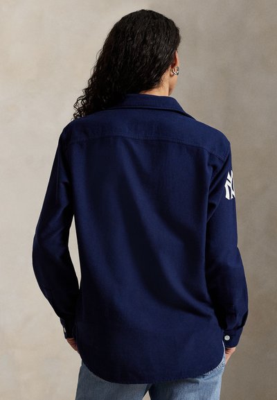 Camisa azul-marinho com botões, com colarinho e mangas longas, apresentando um logotipo branco na manga esquerda. Tecido macio com uma textura suave.