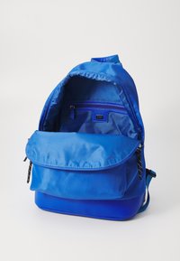 Sac à dos bleu avec un extérieur doux, doté d'une fermeture à rabat, d'une poche zippée intérieure et d'un panneau inférieur texturé. Tissu lisse sur l'ensemble.