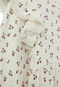 Pamučna haljina s krem pozadinom, koja ima cvjetni uzorak s malim tamnocrvenim višnjama. Rukavi s volanima imaju čipkastu obrubu.