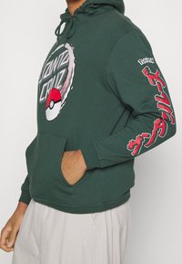 Sudadera verde oscura con los logotipos de Santa Cruz y Poké Ball en el pecho, y texto japonés rojo con la marca Pokémon en la manga.
