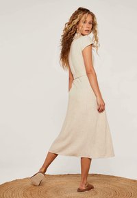 Beiges knie-lang Kleid mit kurzen Ärmeln und tailliertem Schnitt. Hergestellt aus strukturiertem Stoff mit seitlichen Taschen. Getragen mit braunen Sandalen auf einem geflochtenen Teppich.