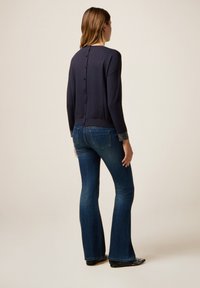 Vrouw in een donkergekleurd, met knopen gesloten, trui met versierde manchet, blauwe flare jeans en gesteelde zwarte schoenen, staande tegen een effen achtergrond.