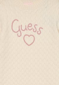 Maglione a trecce crema con il testo ricamato in rosa "Guess" e un cuore. Scollo rotondo e motivo testurizzato su tutta la superficie.