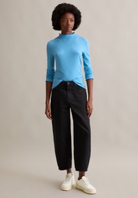 Helles blauer, gerippter langärmeliger Oberteil mit Rüsche am Ausschnitt, kombiniert mit schwarzen, lockeren Hosen und weißen Sneakers. Glatte Textur, lässige Passform.