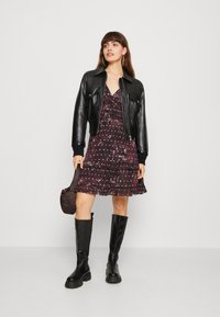 Jaqueta bomber de couro preta sobre um vestido escuro com estampado floral e babados, combinada com botas pretas até ao joelho e uma mala de ombro castanha.