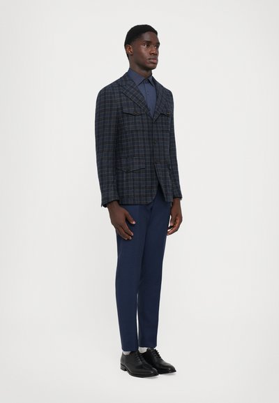 Sacou din țesătură în carouri bleumarin, cu două buzunare frontale, asociat cu o pereche de pantaloni asortați și pantofi negri cu șireturi. Material neted, design structurat.