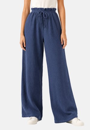 Frau trägt locker sitzende, hoch taillierte marineblaue Hose mit Kordelzug in der Taille, kombiniert mit weißen Sneakers und einem weißen Top.