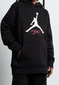 Czarna bluza z kapturem zdobiona białym logo Jumpman i czerwonym napisem „Flight”. Wykonana z miękkiego materiału, posiada kieszeń z przodu oraz ściągacze na mankietach.