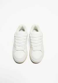 Fehér sportcipők sima szintetikus felsőrésszel, kerek orral és lapos, érdes talppal. Kiugró fűzőkkel és párnázott bokarésszel rendelkezik.