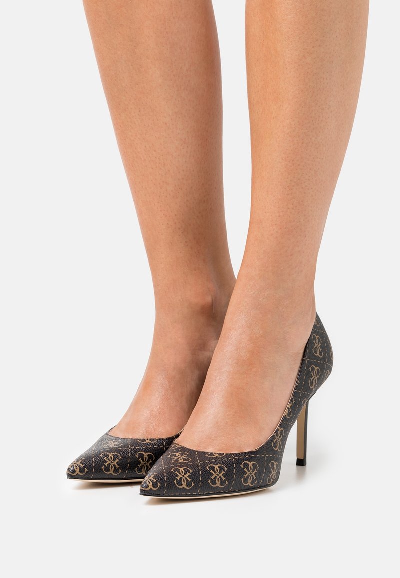 Jambes portant des chaussures noires à talons hauts et bouts pointus avec un motif monogramme beige sur fond blanc.