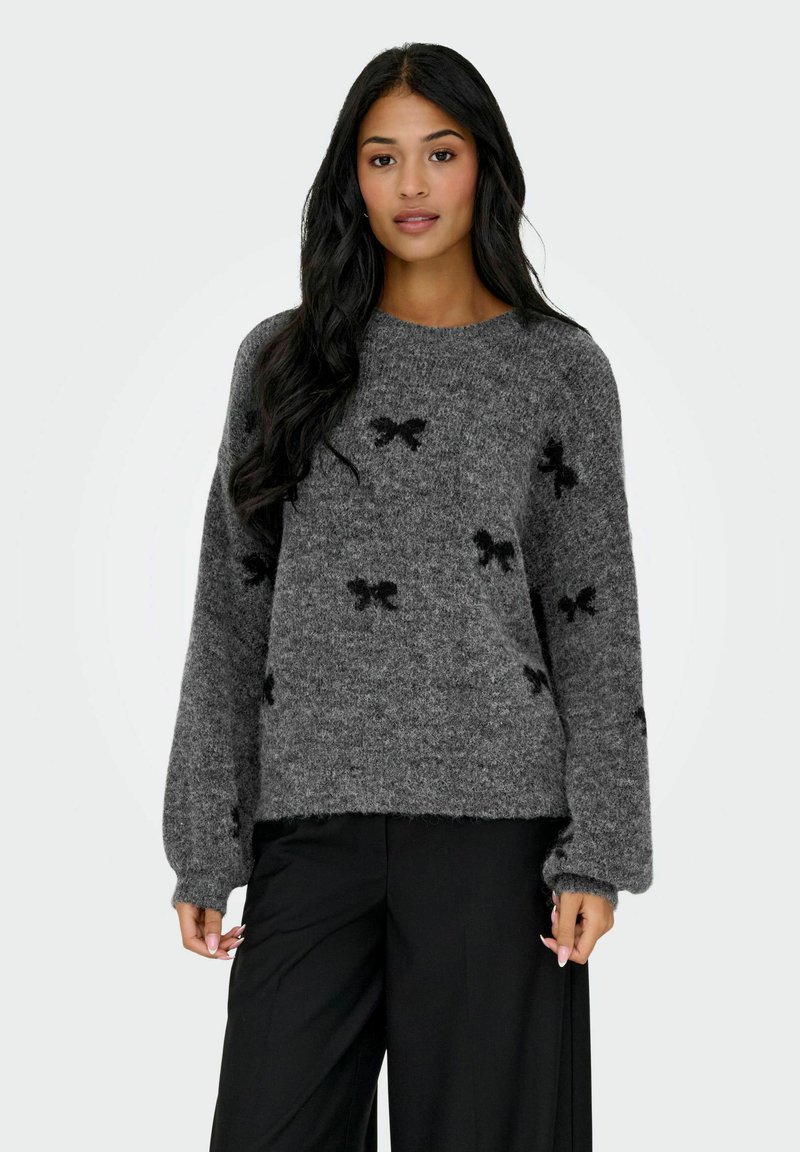 Vrouw met lang zwart haar, gekleed in een grijze fuzzy trui met zwarte strikpatronen en zwarte wijde broek, staande tegen een effen achtergrond.