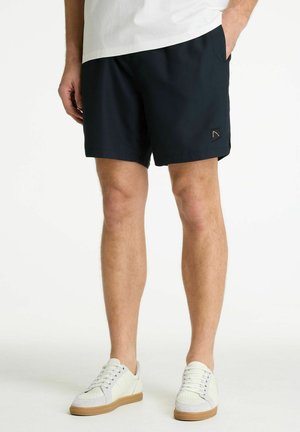 Mees seisab, seljas mustad lühikesed püksid väikese logoga vasakul jalal, valged tossud ja valge särk, mis on pükstesse toppitud.