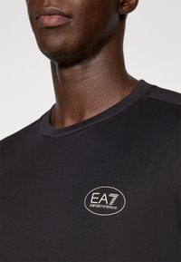 Črna majica s kratkimi rokavi z okroundim izrezom in gladko teksturo, ki na prsih nosi ovalni bel logo "EA7 EMPORIO ARMANI".