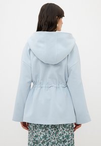 Chaqueta con capucha de color azul claro, con cintura ajustada, mangas largas y una textura de tela suave. La parte trasera presenta una silueta lisa y costuras sutiles.