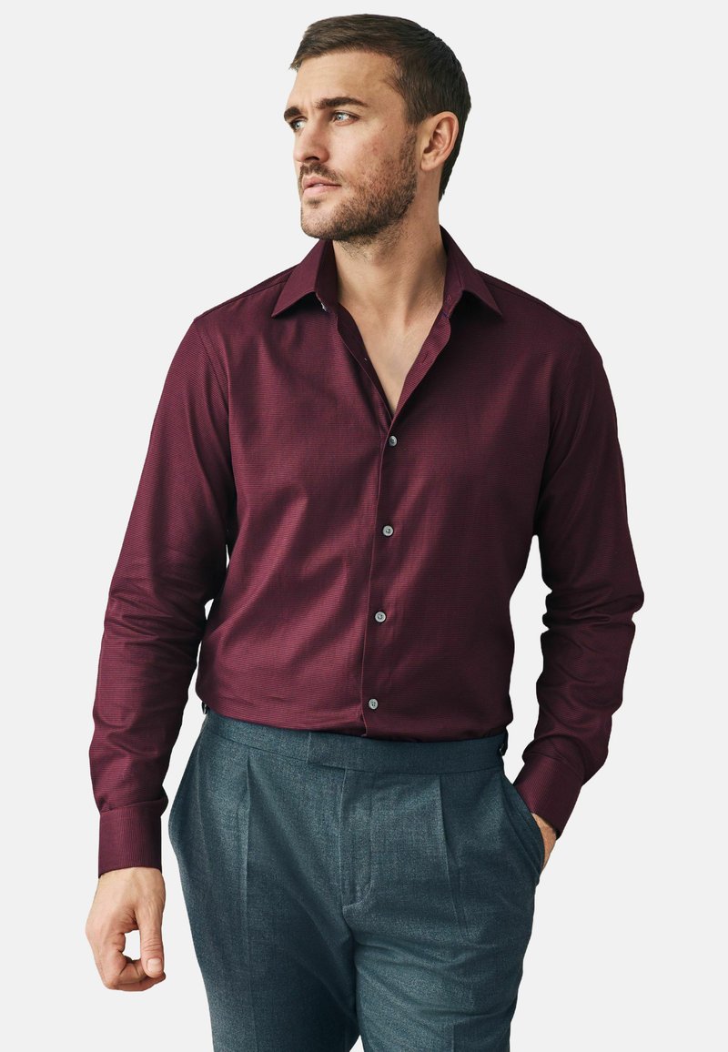 Cămașă burgundy cu nasturi, cu o textură subtilă, având guler și mâneci complete; asortată cu pantaloni gri pliați.