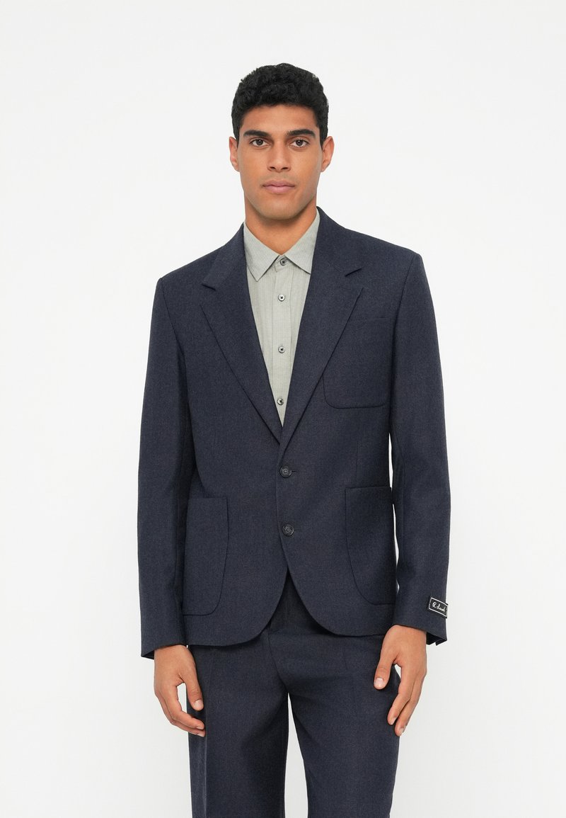 Μπλε μάλλινο σακάκι με δύο μπροστινές τσέπες, κλείσιμο με δύο κουμπιά, γιακάδες με αιχμηρή ακμή και διακριτικό υφασμάτινο φινίρισμα, συνδυασμένο με ανοιχτό γκρι πουκάμισο με γιακά.