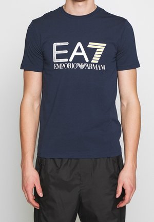 Man draagt een marineblauw T-shirt met het logo "EA7 Emporio Armani" in wit en geel, gecombineerd met een zwarte broek.