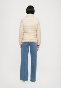 Vrouw die wegkijkt, gekleed in een beige gewatteerde jas, wijde blauwe jeans en witte schoenen tegen een effen lichte achtergrond.
