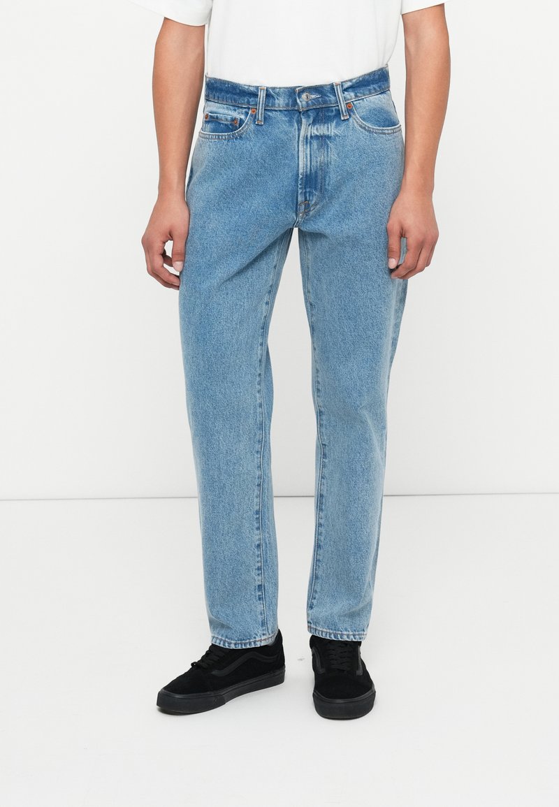 Világoskék farmernadrág laza szabással, öt zsebbel és fémből készült gombos záródással. Fekete fűzős sportcipővel viselve.