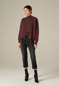 Cardigan bordeaux con bottoni, abbinato a un maglione bordeaux, jeans neri a vita alta e tacchi a stiletto neri. Texture in maglia morbida.