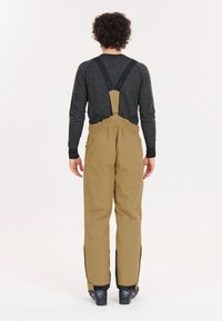Bruine waterdichte skibroek met zijritsen, verstelbare bretels en elastische tailleband, gecombineerd met een donker langomarm shirt.