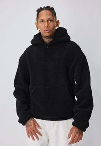 Sweat à capuche en polaire noire avec une coupe décontractée, fermeture éclair, poche avant et matériau texturé. Dispose de manches surdimensionnées et d'un col montant.