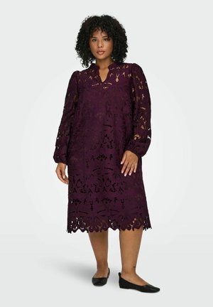 Robe en dentelle violette avec un col haut, des manches longues bouffantes et des motifs découpés floraux avec un ourlet festonné. Portée avec des chaussures plates noires.