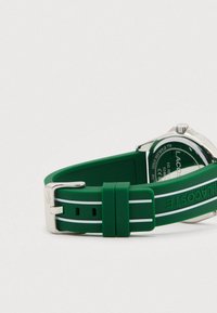 Roheline silikoonist kella rihm valgete triipudega ja hõbedane pandla, osaliselt nähtav roostevabast terasest kella tagakülg Lacoste brändimisega.