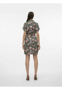 Vestito floreale verde con fiori rosa e bianchi, maniche corte, vita con cintura e lunghezza fino al ginocchio. Sandali con tacco completano il look.