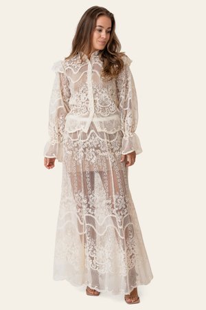 Naine, kes kannab pikka, läbipaistvast valgest pitsist kleiti lillemustriga, esiküljel nööpidega ja pikkade volangiliste varrukatega, seisab ühevärvilise tausta ees.