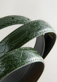 Green belt made of textured leather with a snake scale pattern, featuring contrasting stitching and smooth edges. Circular shape.