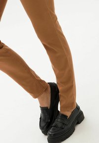 Pantalones marrones de corte slim con una textura suave, combinados con mocasines negros con un acento con flecos y suelas gruesas.