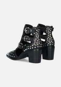 Botas de tobillo negras con tacón alto, que presentan tres hebillas ajustables, detalles de tachuelas y lados abiertos. Fabricadas en cuero con un tacón grueso.