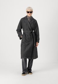 Abrigo gris de espiga de doble botonadura con cinturón, mangas largas y bolsillos de parche. El modelo lleva gafas de sol negras y un cuello de alto gris.