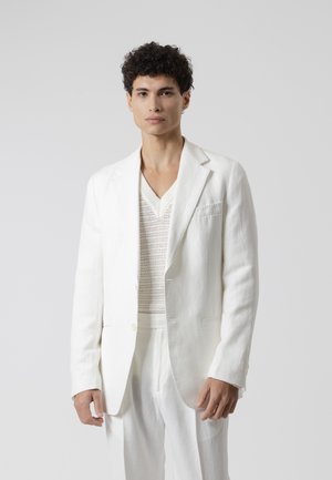 Joven con cabello rizado que viste un conjunto totalmente blanco, que incluye una chaqueta, un suéter de punto y pantalones, de pie frente a un fondo claro y liso.