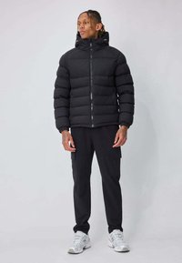 Veste matelassée noire avec fermeture éclair devant, col montant et texture matelassée horizontale, associée à un pantalon cargo noir et des baskets blanches.