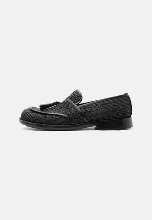 Mocassin tissé noir avec détail pompon, petit talon et bout arrondi, présenté en profil latéral sur fond blanc.