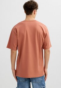 Joven con cabello corto y rizado, vistiendo una camiseta oversize lisa de color rosa polvo y jeans de mezclilla azul, de pie y de espaldas a la cámara.