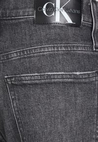 Harmaat farkut, joissa on näkyvä etiketti, joka esittelee "Calvin Klein Jeans" -merkin. Korostuksia ovat ompelu yksityiskohdat ja useita taskuja.